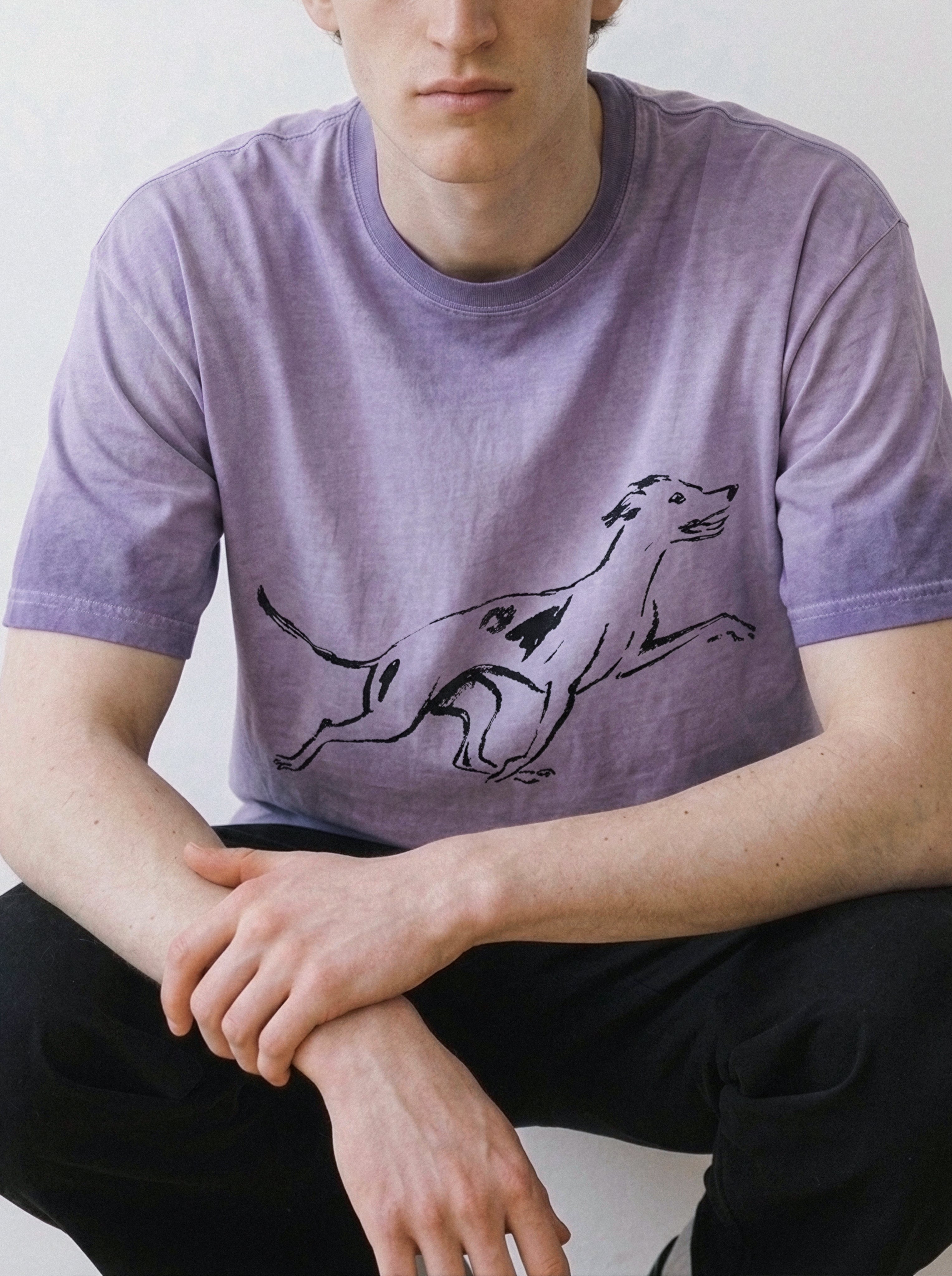 A male model leaning forward, showcasing the relaxed drape of an ombre violet t-shirt highlighting a pure line art running dog graphic.