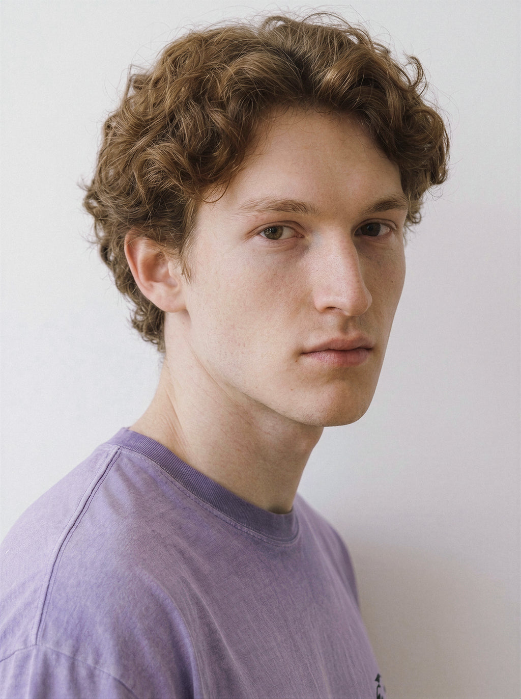A young man with curly light brown hair wearing a light purple crewneck t-shirt, photographed in a close-up portrait against a plain off-white background.