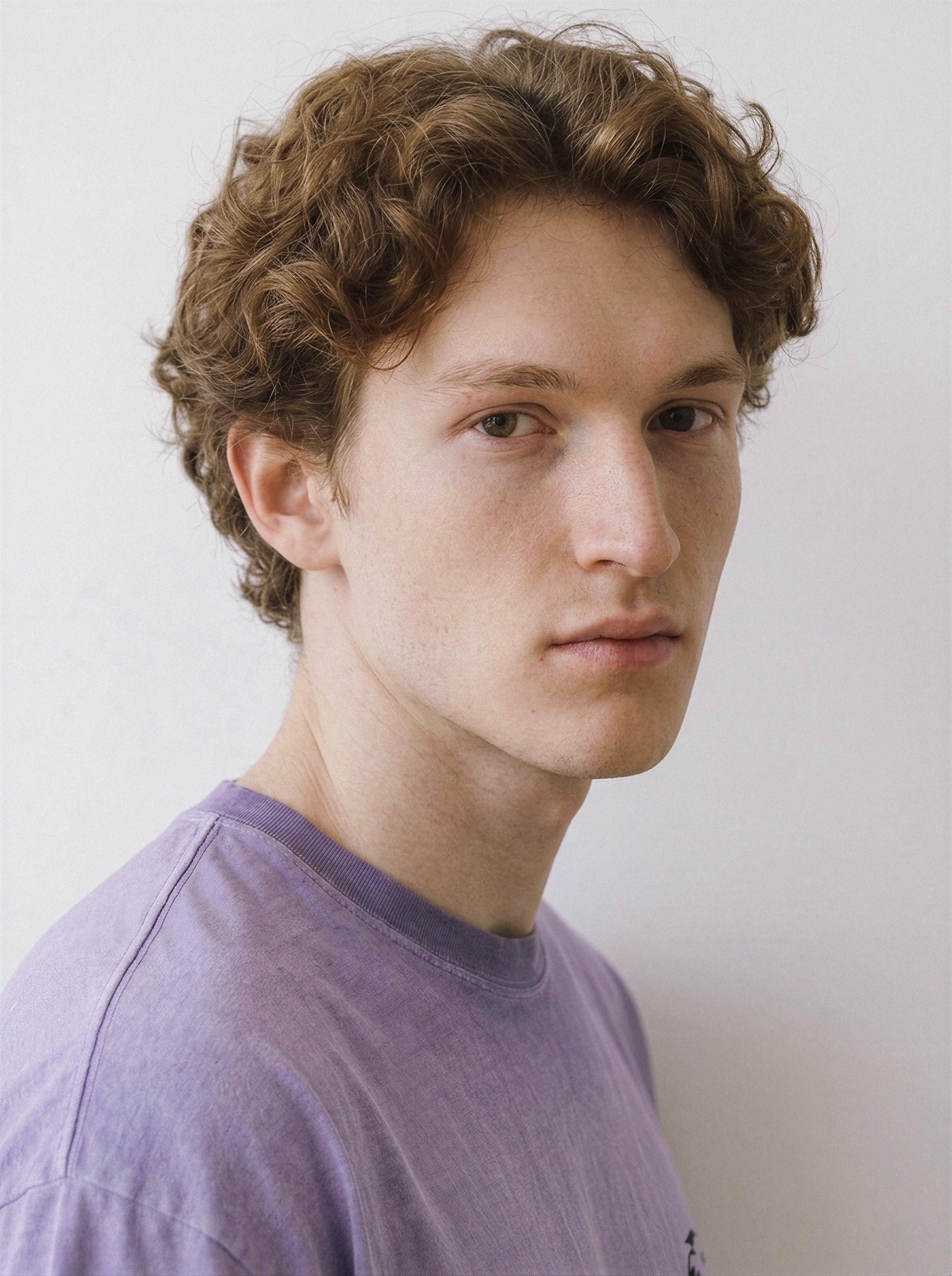 A young man with curly light brown hair wearing a light purple crewneck t-shirt, photographed in a close-up portrait against a plain off-white background.