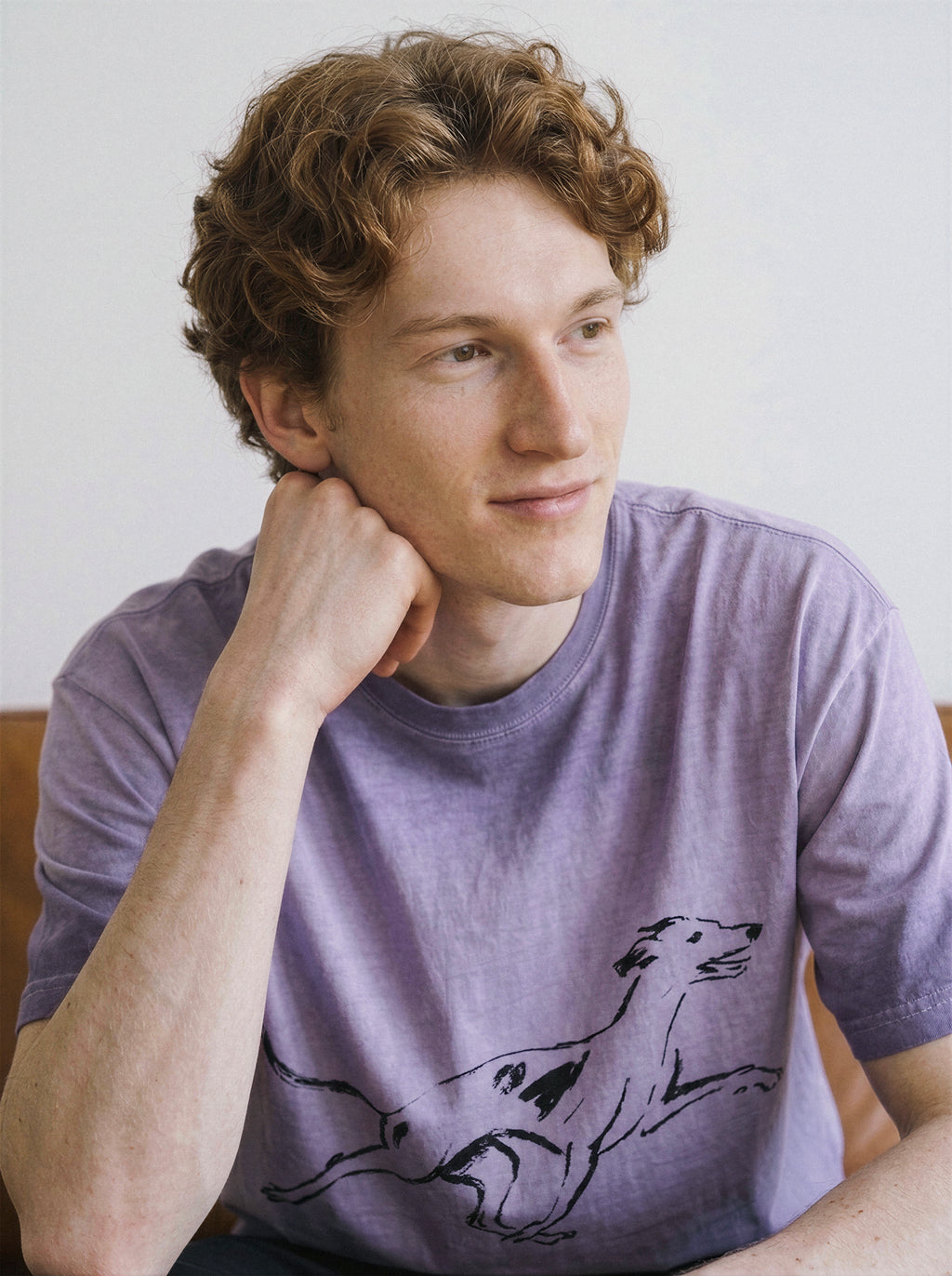 Model sitting and resting his hand on his face, wearing a relaxed ombre violet graphic tee featuring a minimalist black running dog design.