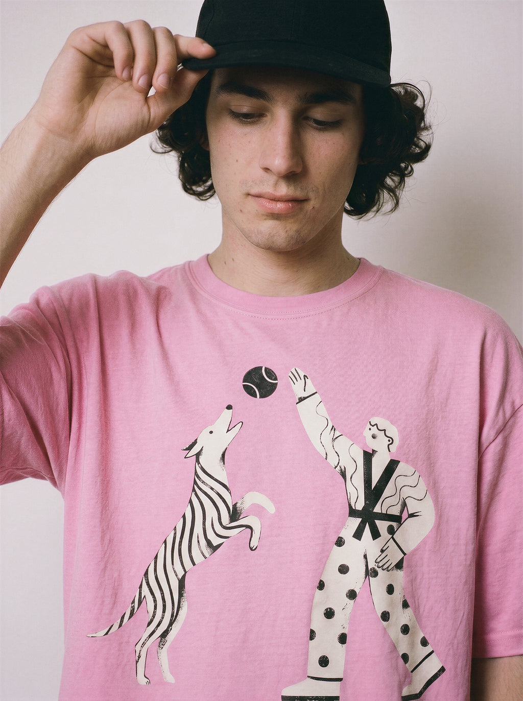 A male model adjusting his cap wearing an aura pink playing fetch ironic graphic tee, capturing the quirky illusion of control design and relaxed fit.