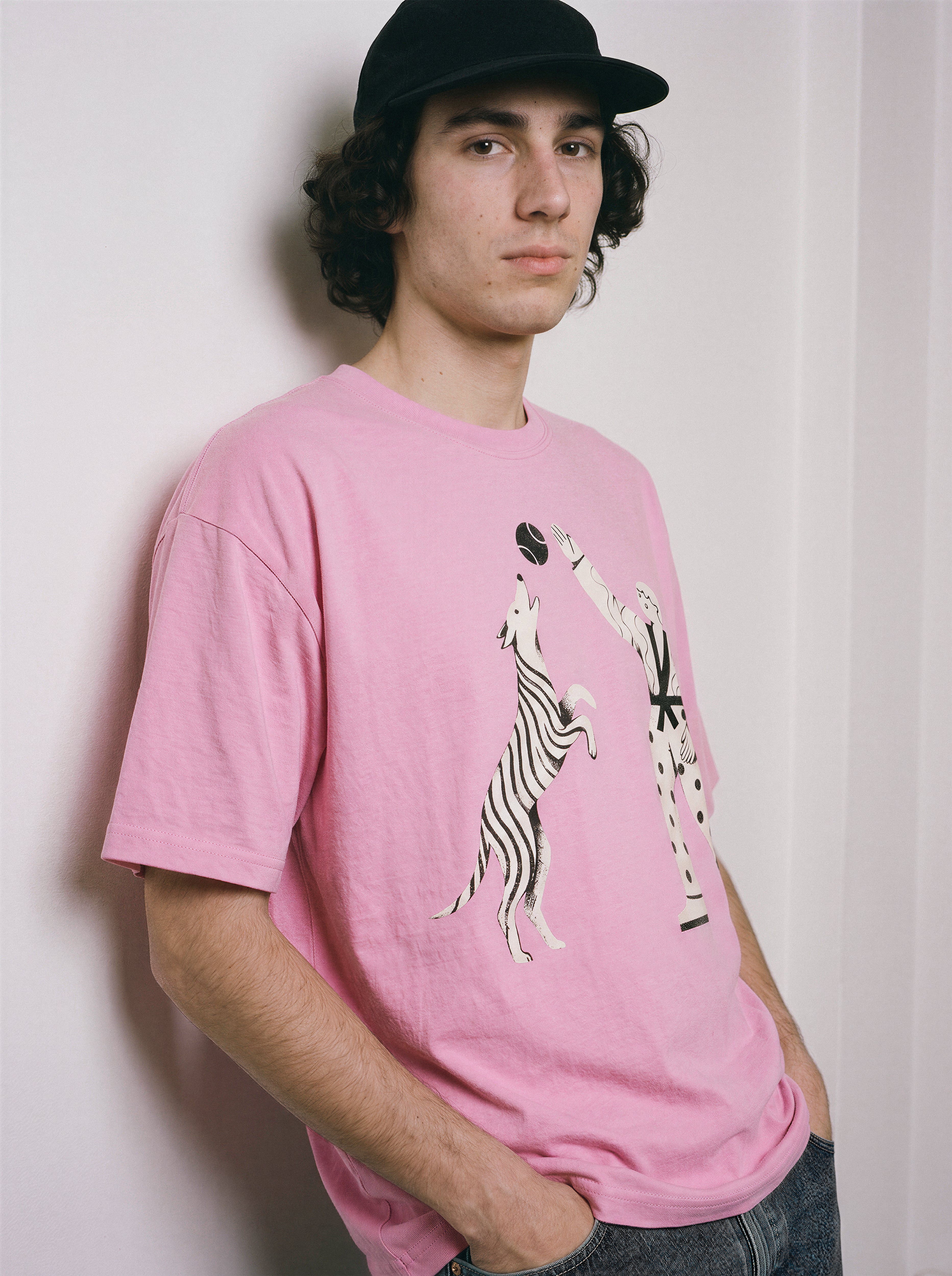 A male model leaning against a wall wearing an aura pink playing fetch ironic graphic tee, demonstrating the casual drape and sarcastic meme aesthetic.