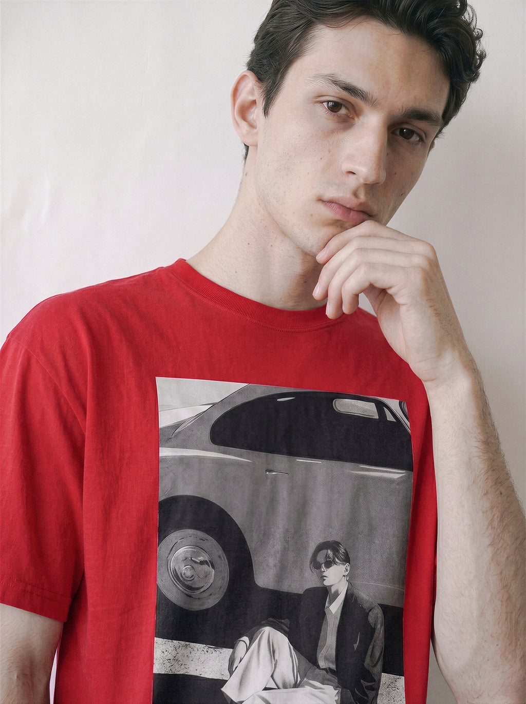 A man wearing a red oversized t-shirt with a 90s retro cinematic photography print on the front showing a vintage car and a seated figure, posed against a plain off-white background.
