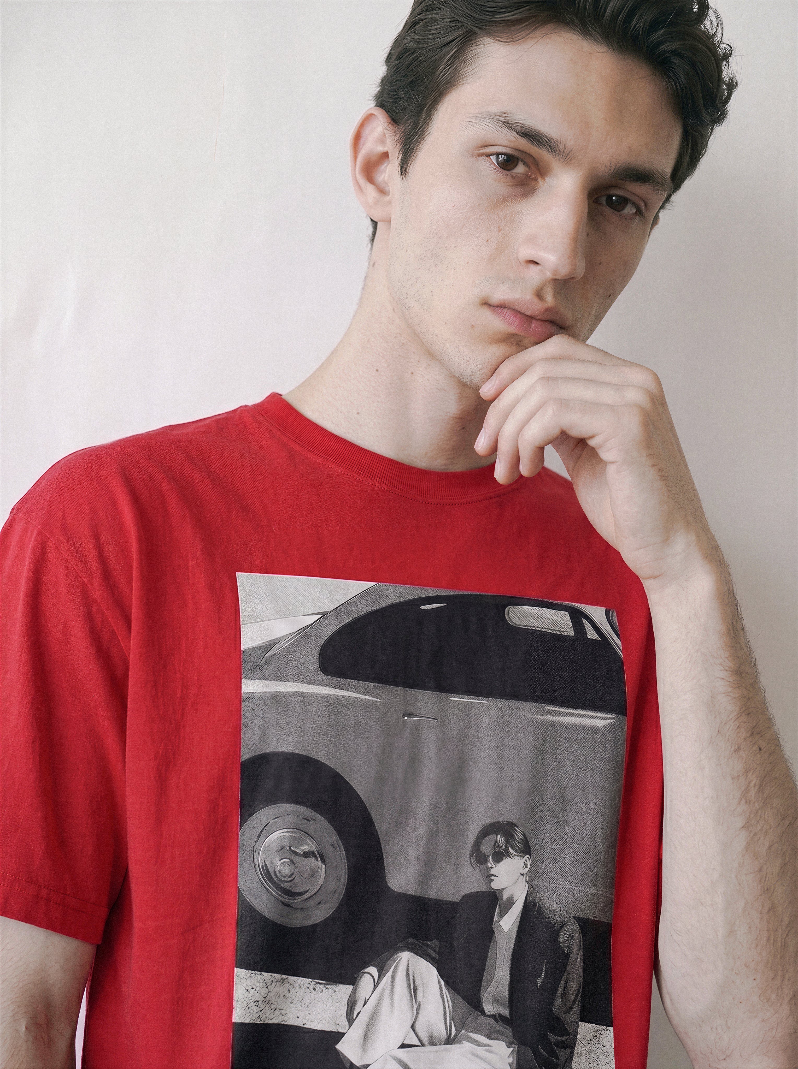 A man wearing a red oversized t-shirt with a 90s retro cinematic photography print on the front showing a vintage car and a seated figure, posed against a plain off-white background.