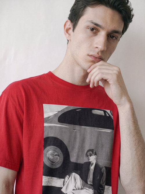 A man wearing a red oversized t-shirt with a 90s retro cinematic photography print on the front showing a vintage car and a seated figure, posed against a plain off-white background.