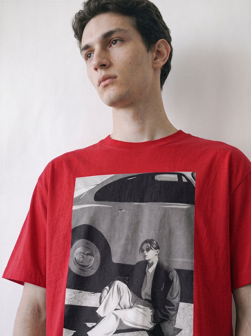 A man wearing a red crewneck t-shirt,clearly displaying the 90s retro cinematic black and white photography print of a vintage car and a seated figure.