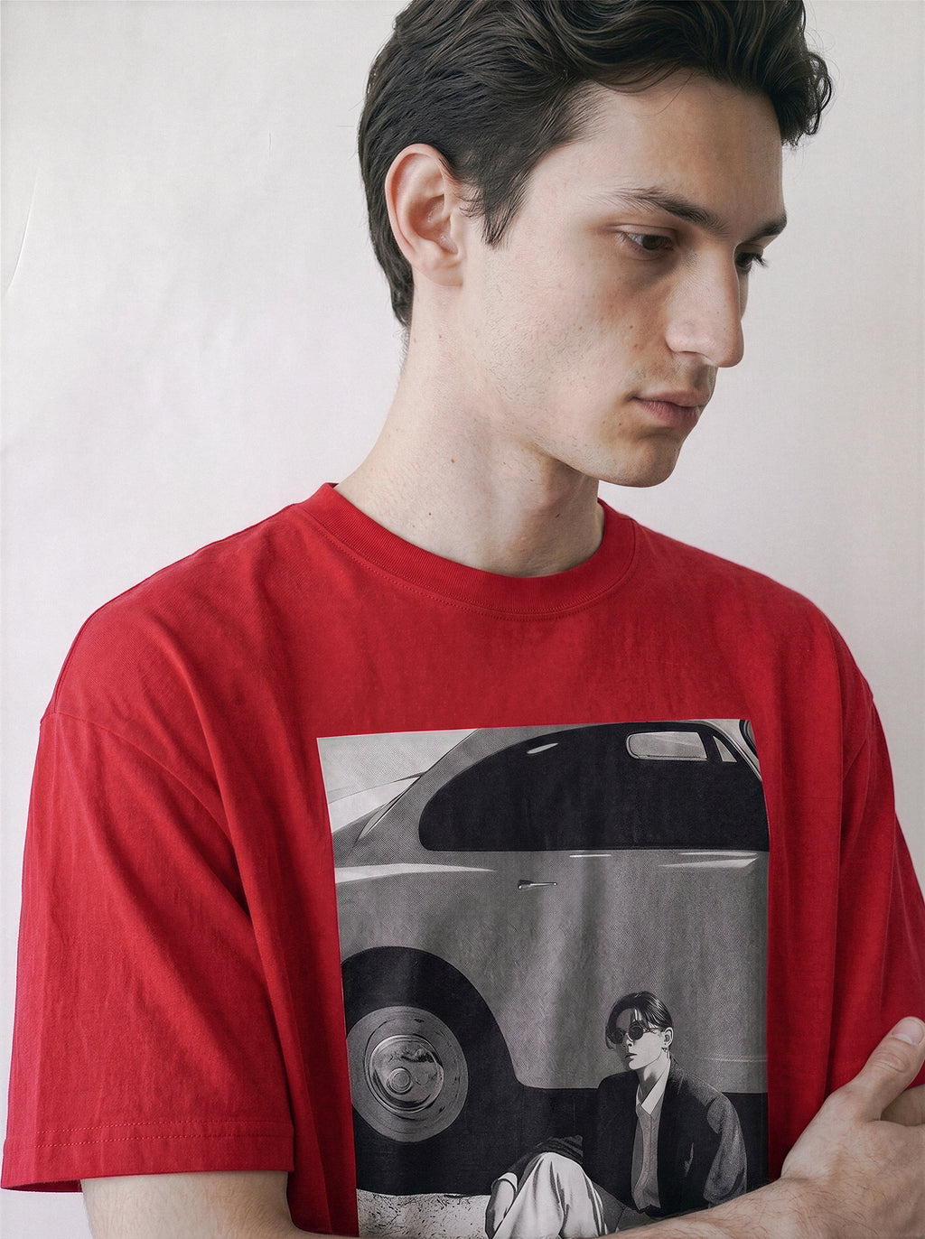 Detailed view of a man looking down, wearing a red oversized indie streetwear t-shirt with a vintage black and white photography print on the front depicting a vintage car and a seated figure, set against a plain off-white background.