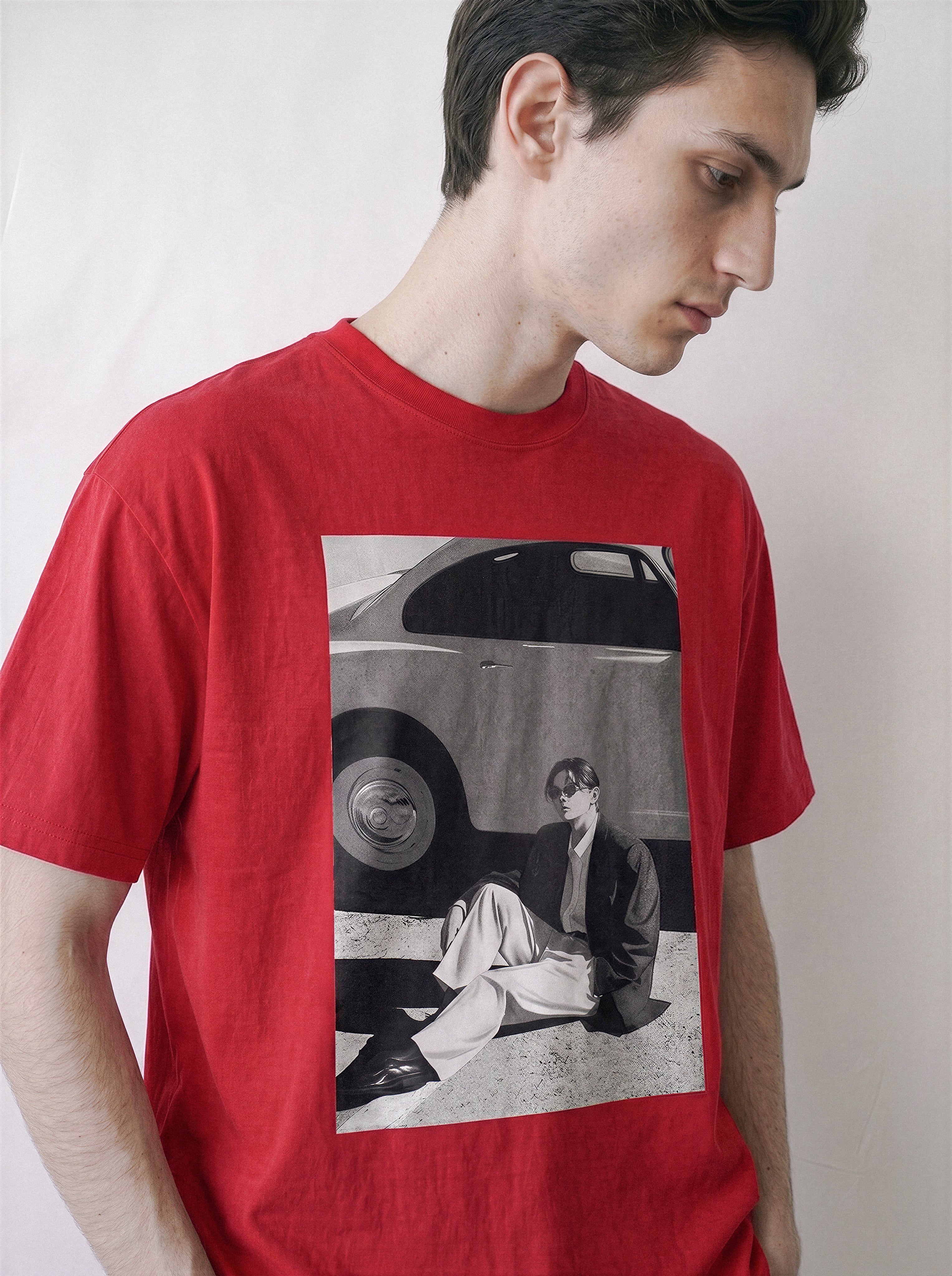 A man wearing a red oversized t-shirt with a 90s retro cinematic black and white photography print depicting a vintage car and a seated figure.