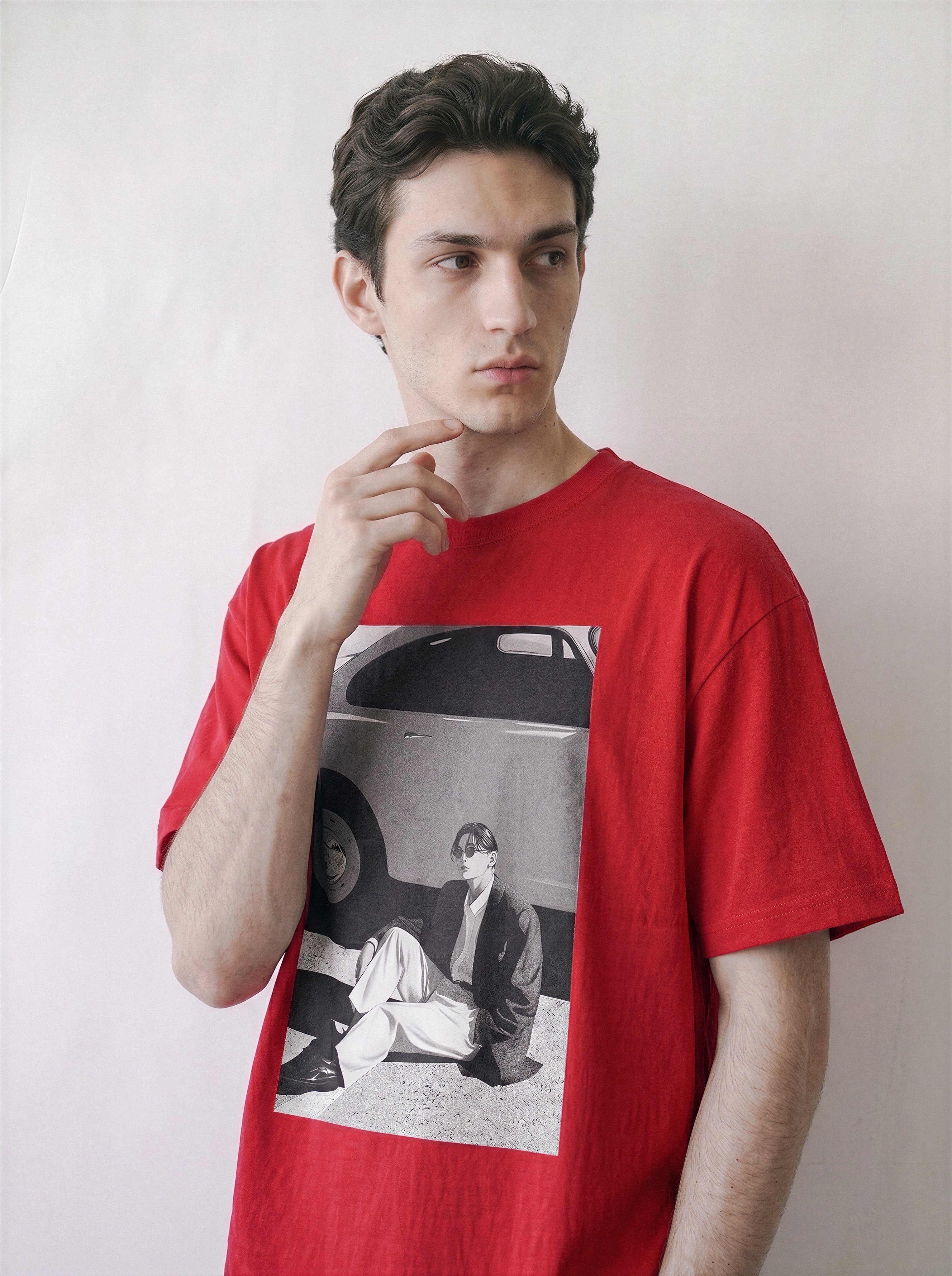 A male model posing with his hand near his neck, wearing a red oversized t-shirt featuring a 90s retro cinematic black and white photography print of a vintage car and a seated figure on the front, posed against a plain off-white wall.