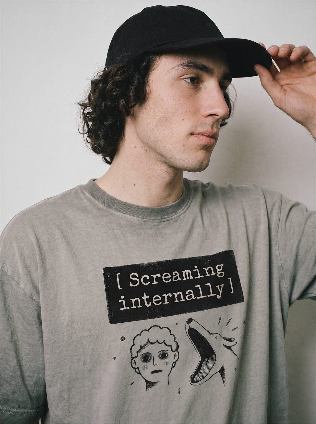 A male model adjusting his cap wearing an ombre grey screaming internally ironic graphic tee, showcasing the sarcastic blank face graphic and Gen Z dry humor vibe.