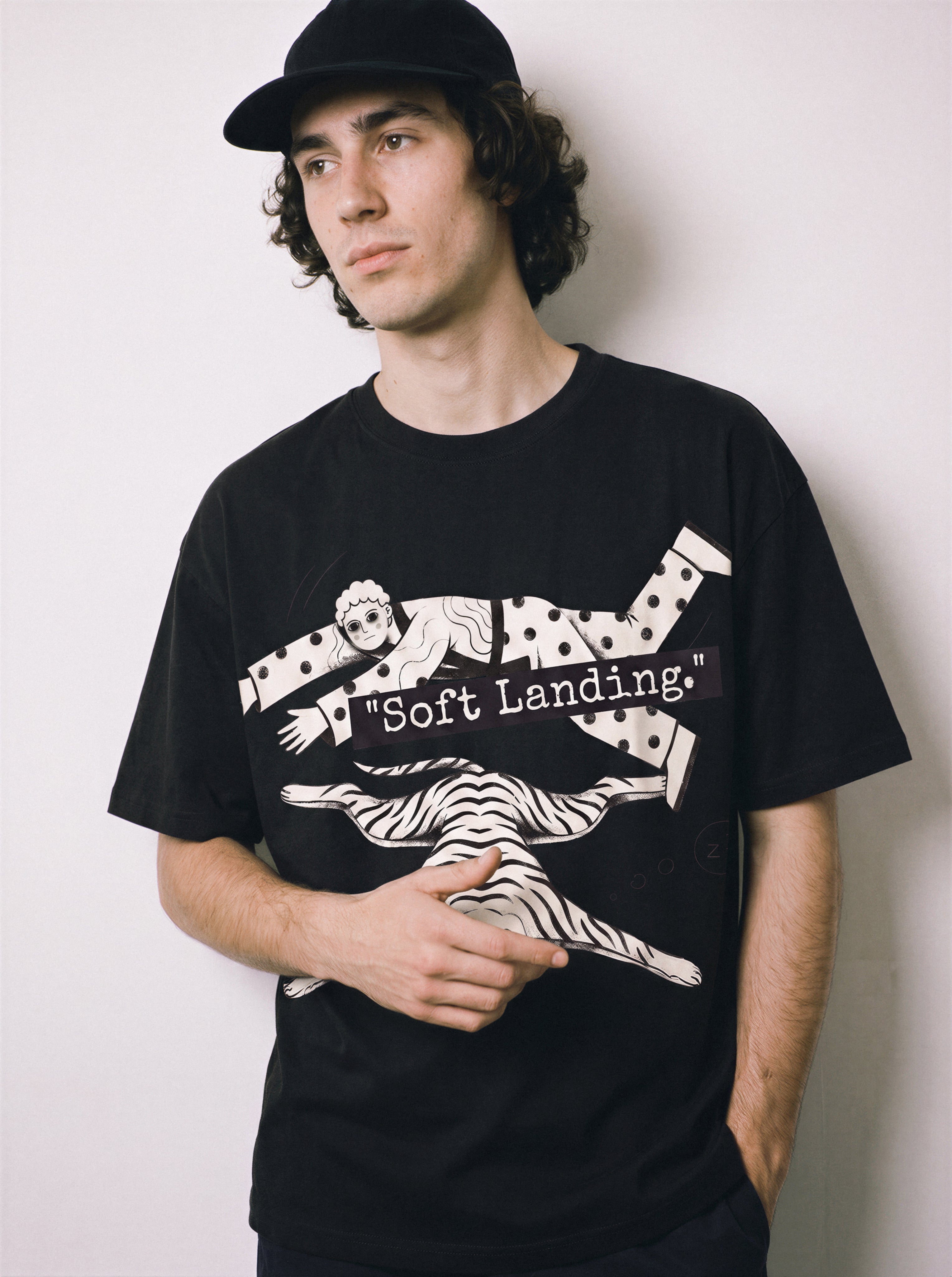 A close-up of a male model holding his shirt hem wearing a black soft landing line art graphic tee, showcasing the person and dog safely hitting the floor graphic.