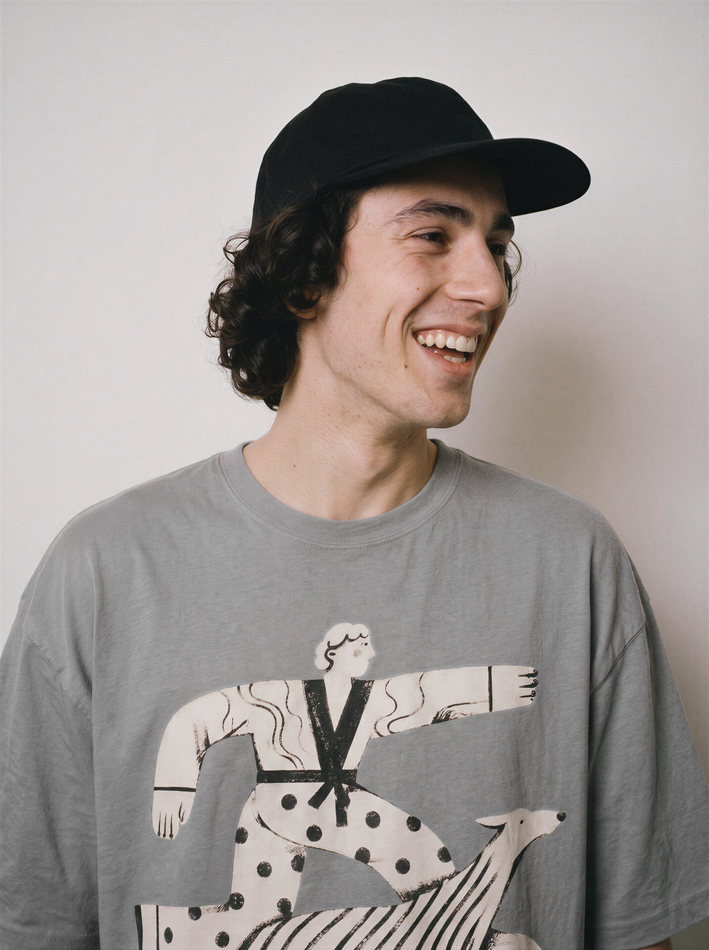 A profile portrait of a smiling male model wearing a gray striding away line art graphic tee, focusing on the detached streetwear style and lighthearted mood.