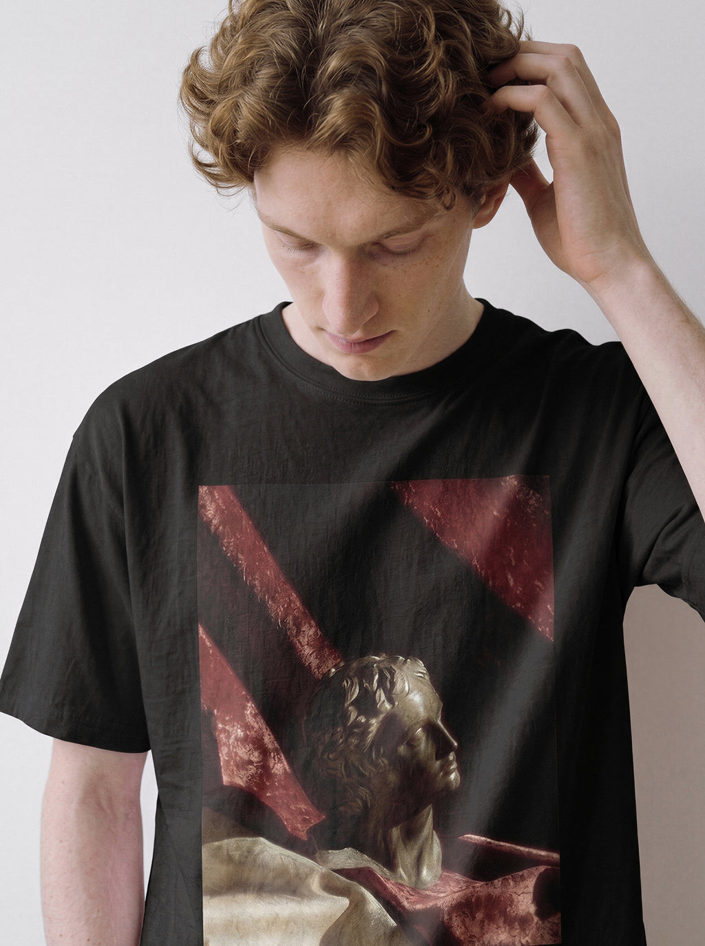 A male model with his head bowed and hand in his hair wearing a black surreal classical bust graphic tee, showcasing the still life apparel aesthetic and high-contrast red drape detail.