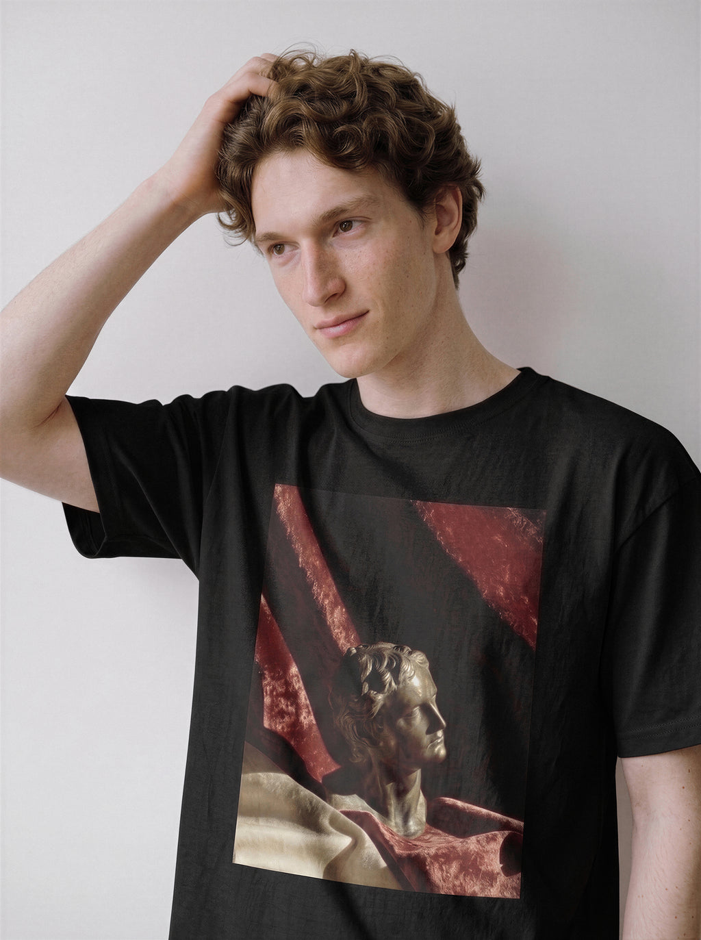 A male model with his hand in his hair looking down wearing a black surreal classical bust graphic tee, showcasing the timeless stone sculpture print and relaxed boxy fit.