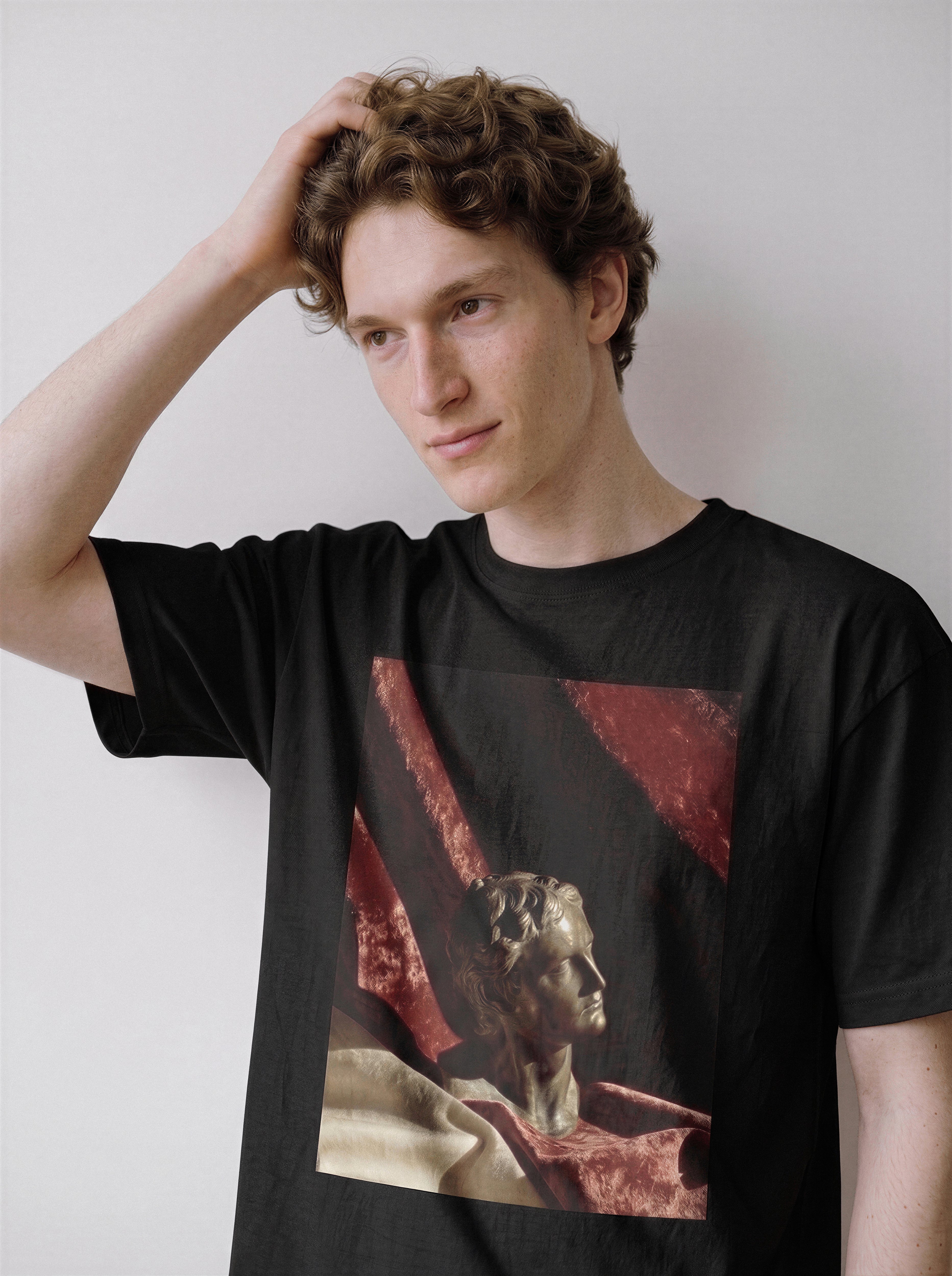 A male model with his hand in his hair looking down wearing a black surreal classical bust graphic tee, showcasing the timeless stone sculpture print and relaxed boxy fit.