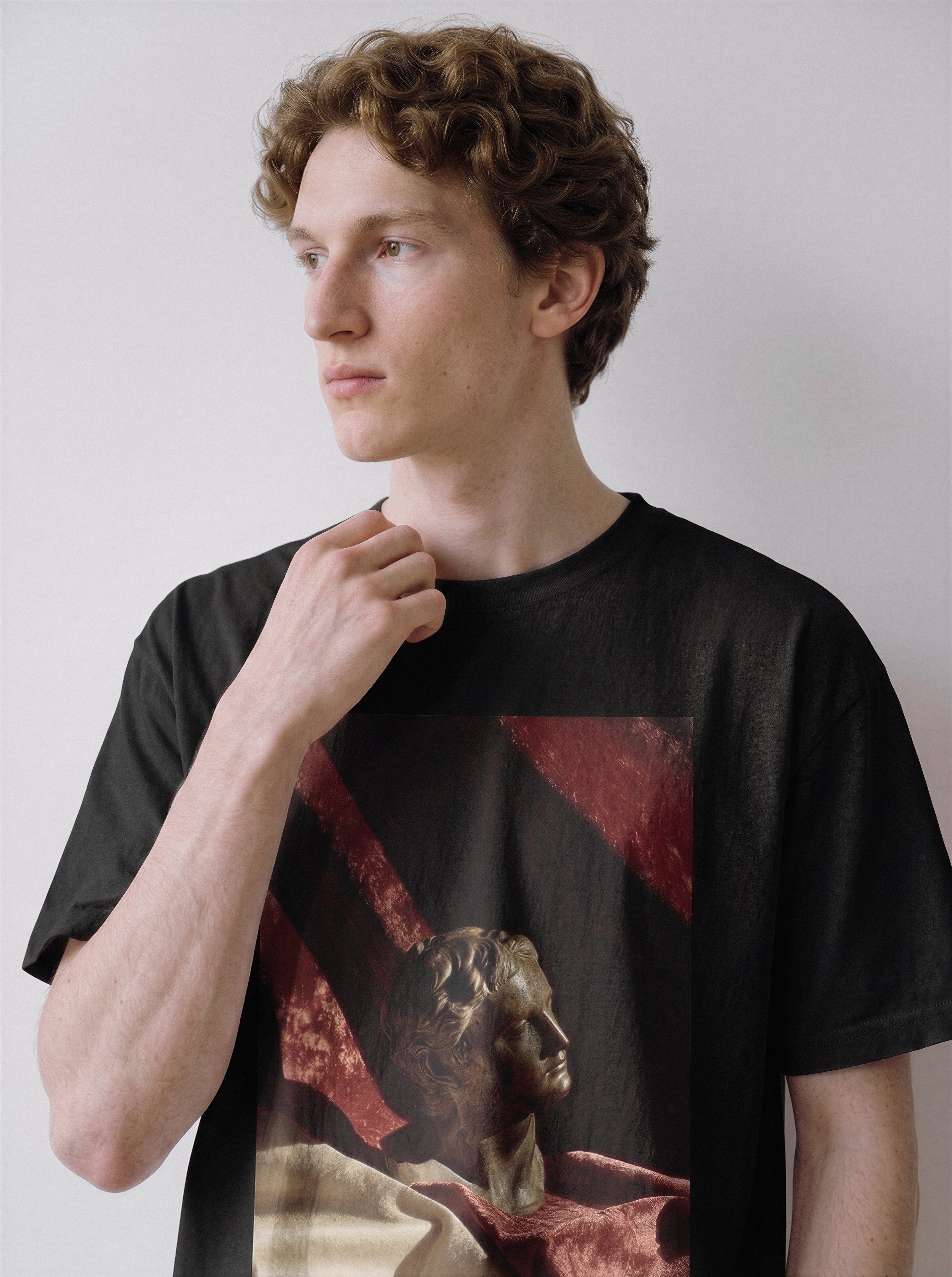 A male model with his hand near his collar looking left wearing a black surreal classical bust graphic tee, focusing on the classical bust print details and timeless visual design.