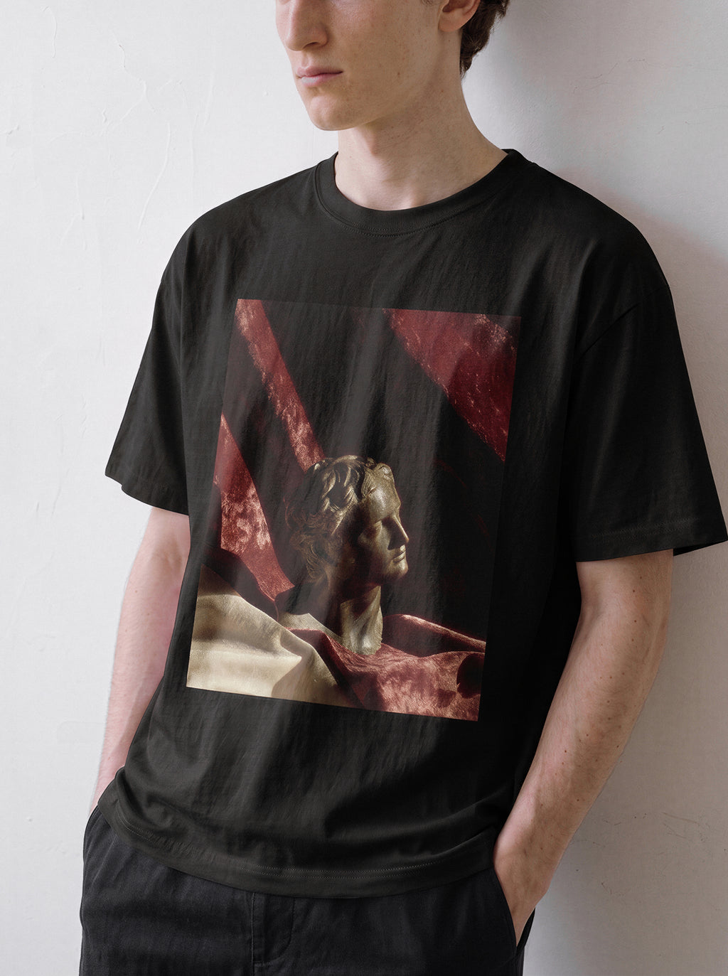 A male model looking down and to the right with hands in pockets wearing a black surreal classical bust graphic tee, displaying the museum core apparel style and objective artistic elements.