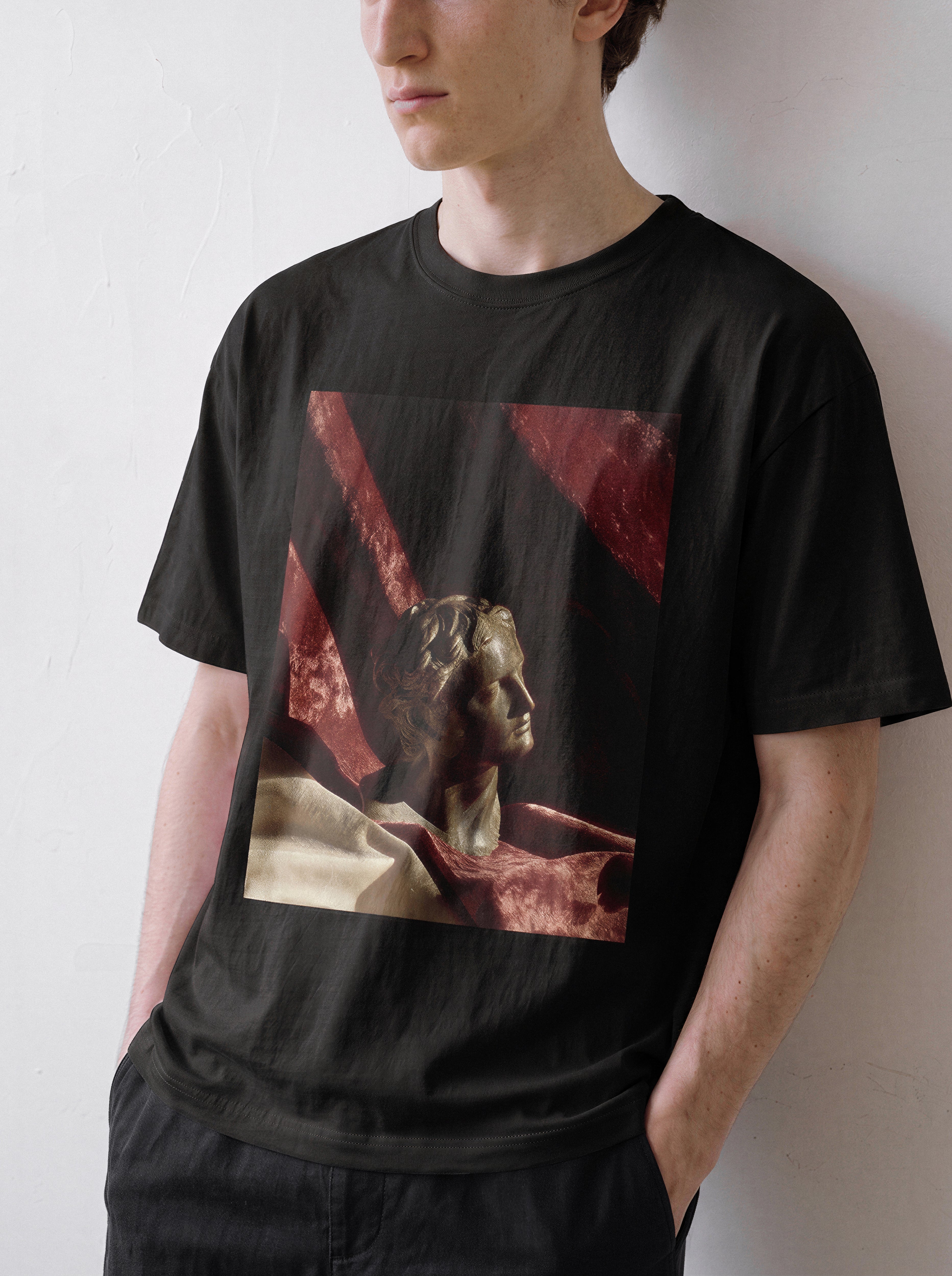 A male model looking down and to the right with hands in pockets wearing a black surreal classical bust graphic tee, displaying the museum core apparel style and objective artistic elements.