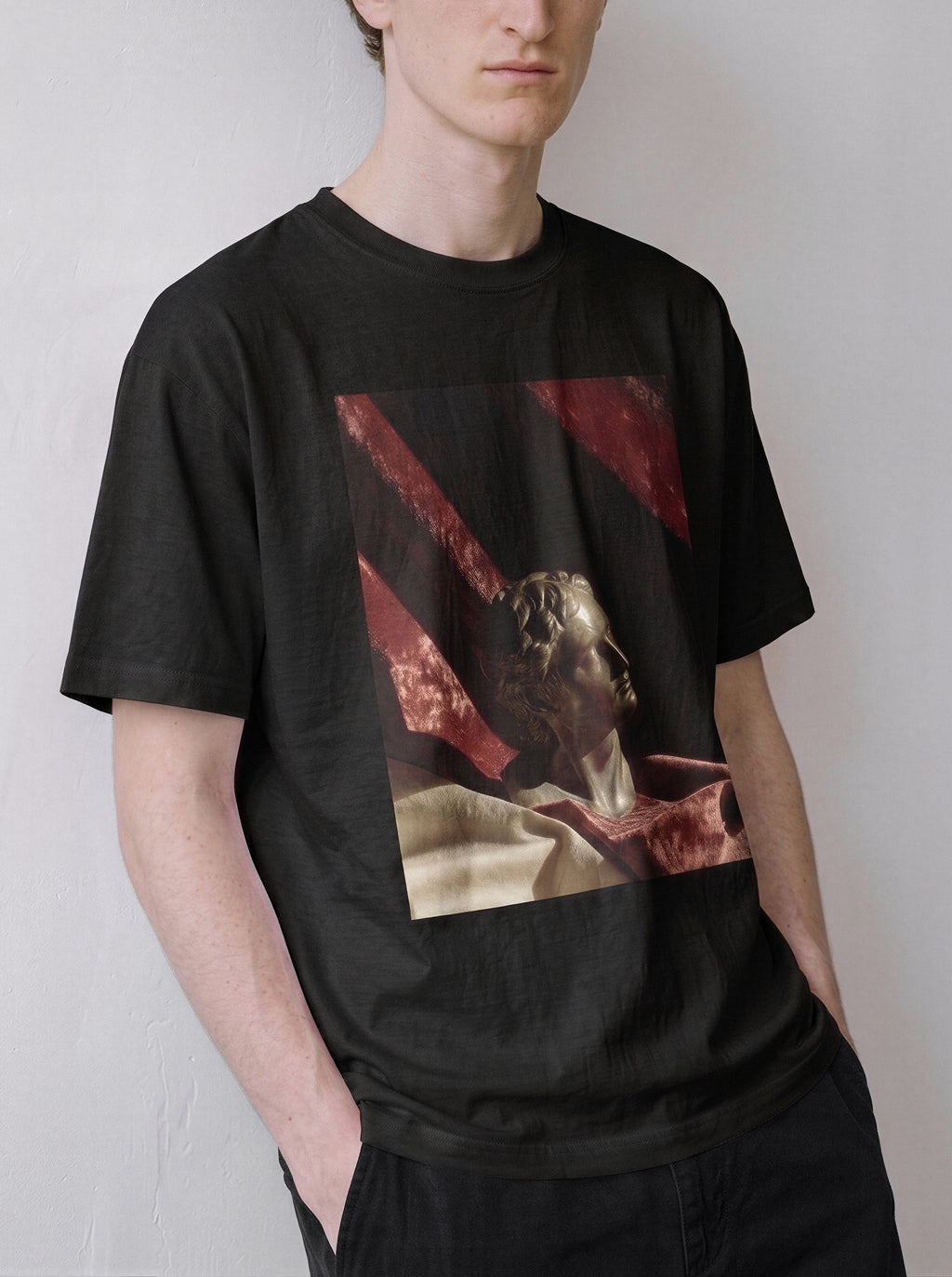 A male model looking to the right with hands in pockets wearing a black surreal classical bust graphic tee, highlighting the classical art streetwear vibe and surreal still life aesthetic.