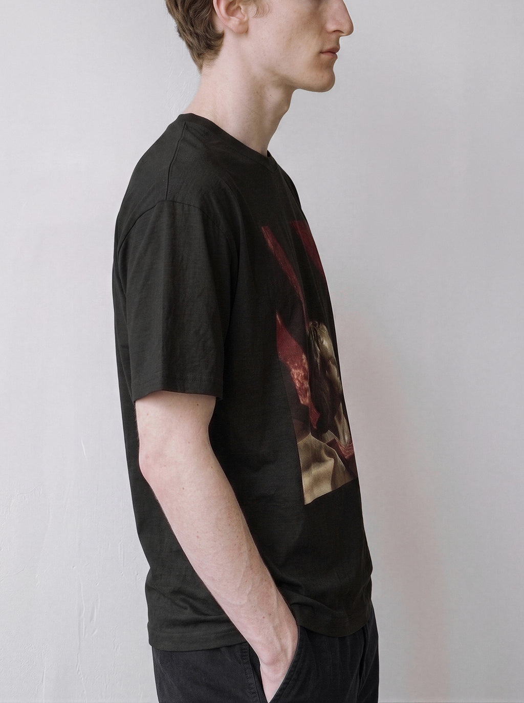 A side profile of a male model standing wearing a black surreal classical bust graphic tee, emphasizing the draped stone statue graphic and casual drape.