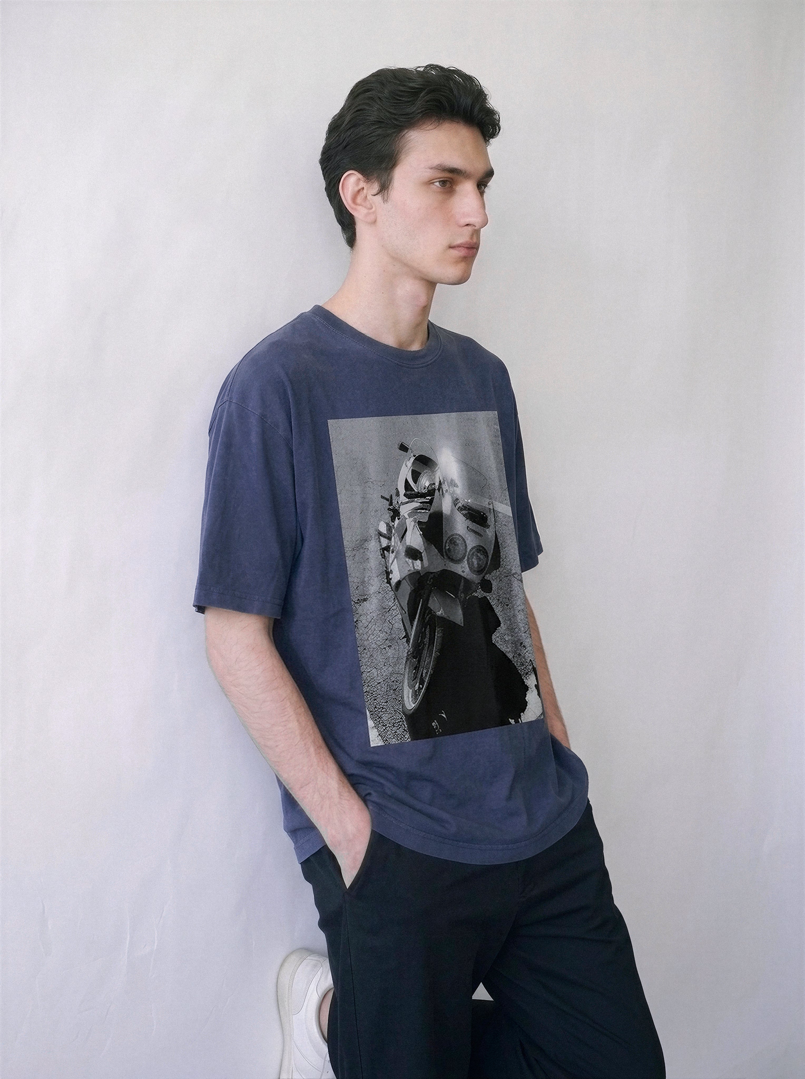 A male model leaning against a wall wearing a vintage navy 90s monochrome motorcycle photography tee, showcasing the relaxed boxy fit and retro motorcore streetwear vibe.