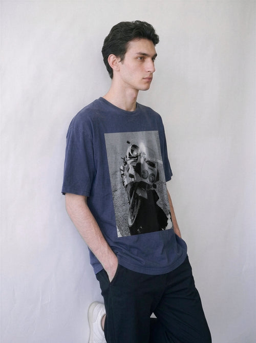 A male model leaning against a wall wearing a vintage navy 90s monochrome motorcycle photography tee, showcasing the relaxed boxy fit and retro motorcore streetwear vibe.