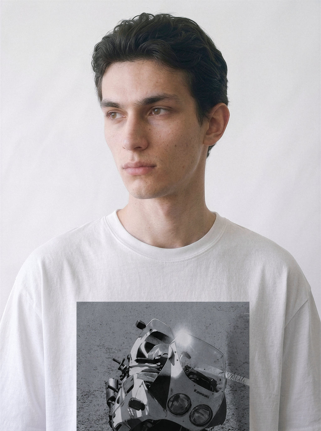 A close-up of a male model looking aside wearing a white 90s monochrome motorcycle photography tee, focusing on the gritty monochrome highway beast print details.