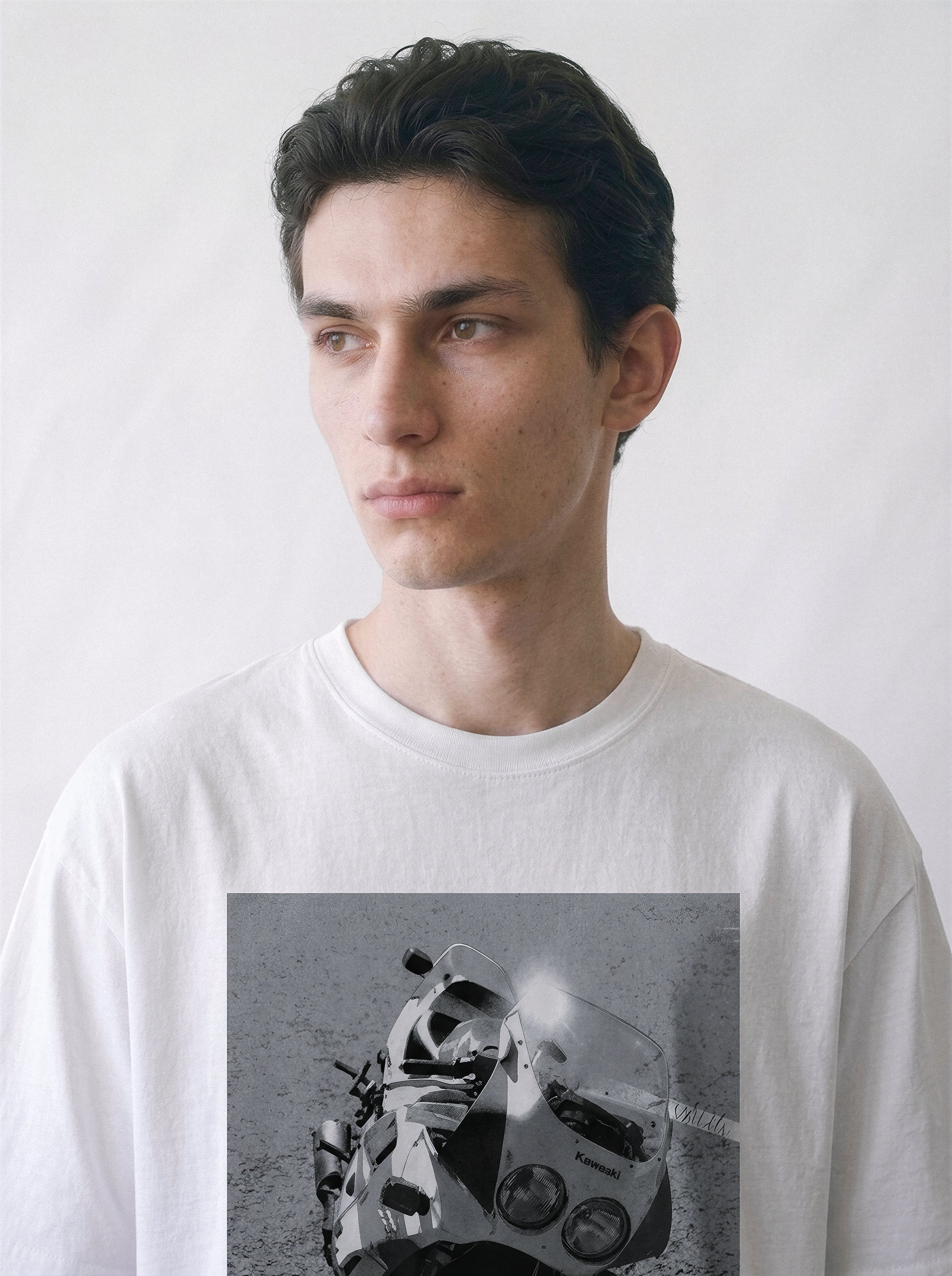 A close-up of a male model looking aside wearing a white 90s monochrome motorcycle photography tee, focusing on the gritty monochrome highway beast print details.