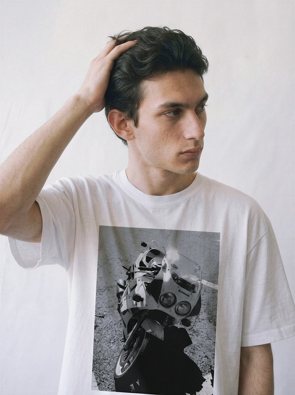 A male model with his hand in his hair wearing a white 90s monochrome motorcycle photography tee, capturing the vintage motorcore apparel style.