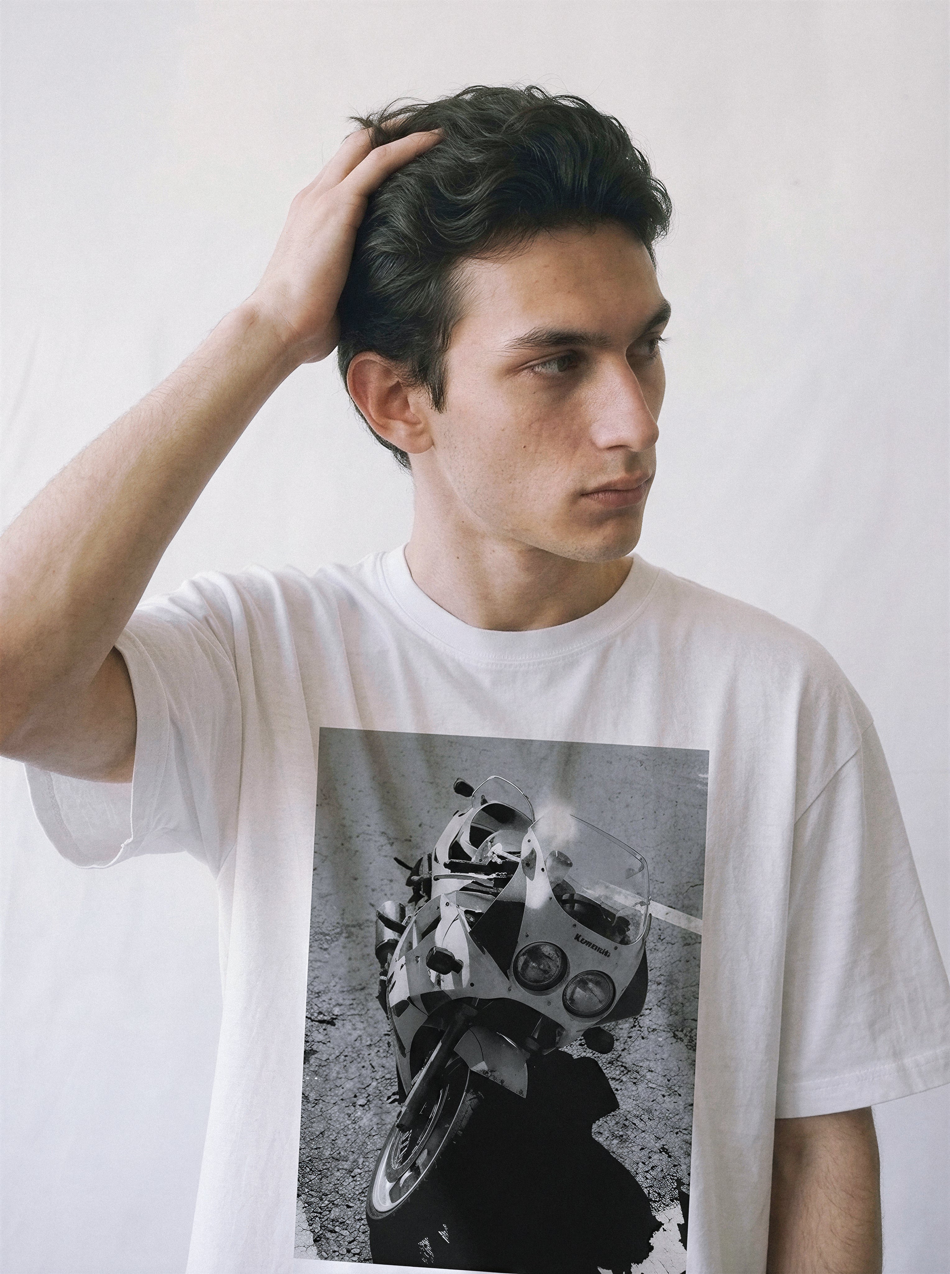 A male model with his hand in his hair wearing a white 90s monochrome motorcycle photography tee, capturing the vintage motorcore apparel style.