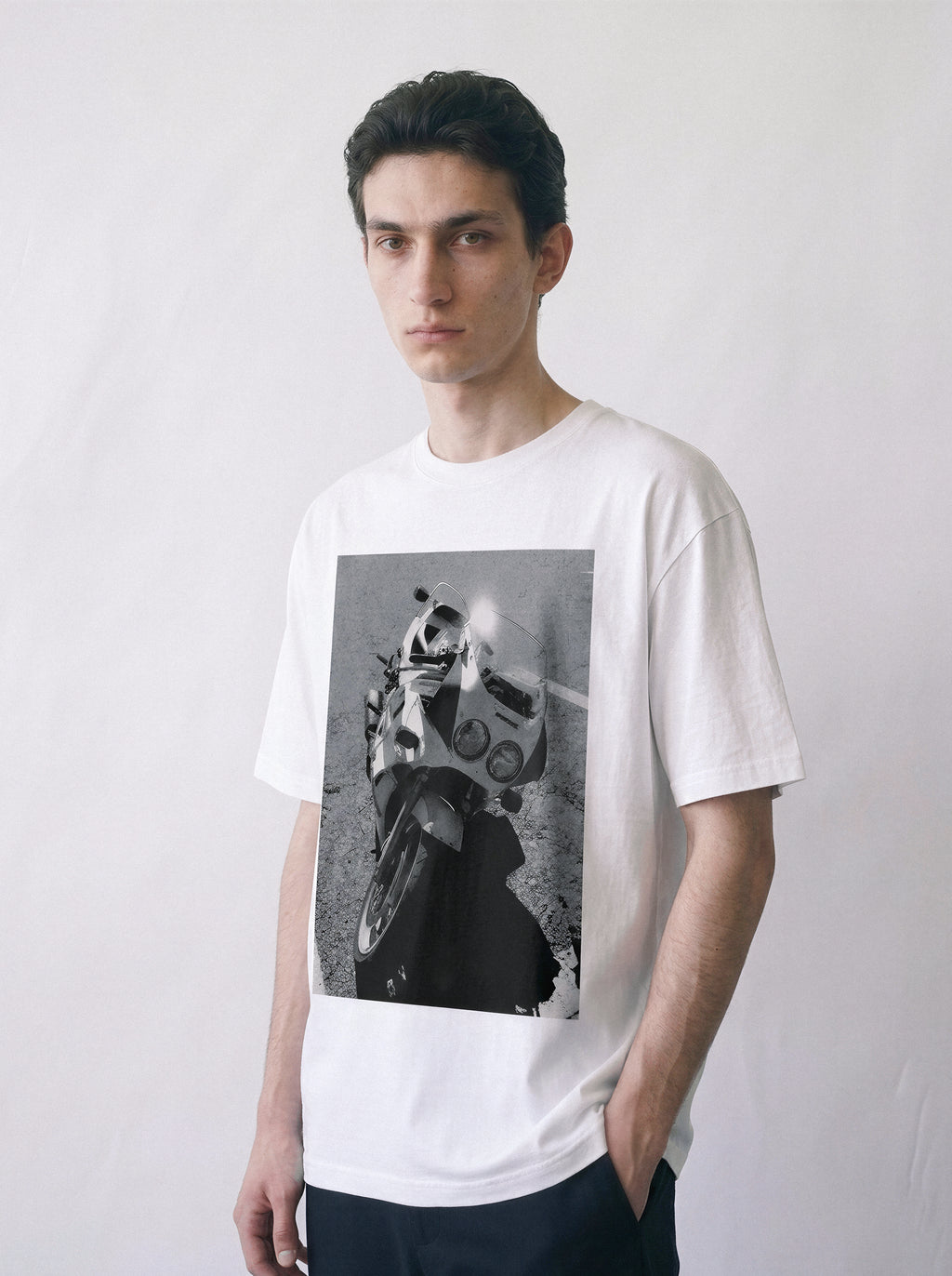 A front view of a male model wearing a white 90s monochrome motorcycle photography tee, displaying the high-contrast mechanical archive graphic.