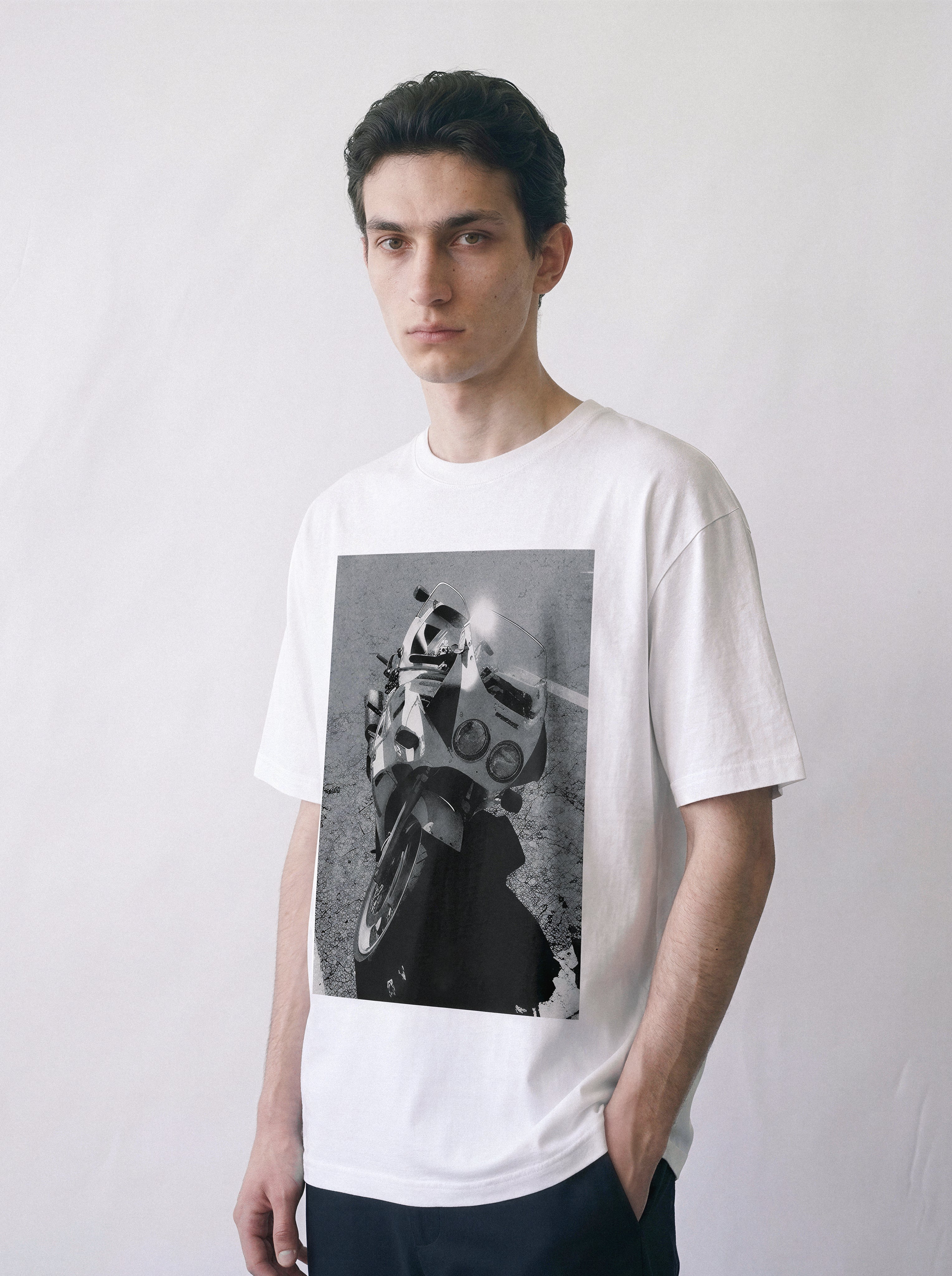 A front view of a male model wearing a white 90s monochrome motorcycle photography tee, displaying the high-contrast mechanical archive graphic.