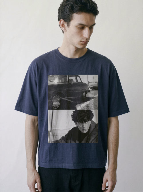 A man wearing a navy blue oversized graphic t-shirt with a black and white collage print of a vintage car and a portrait, set against a plain light gray background.