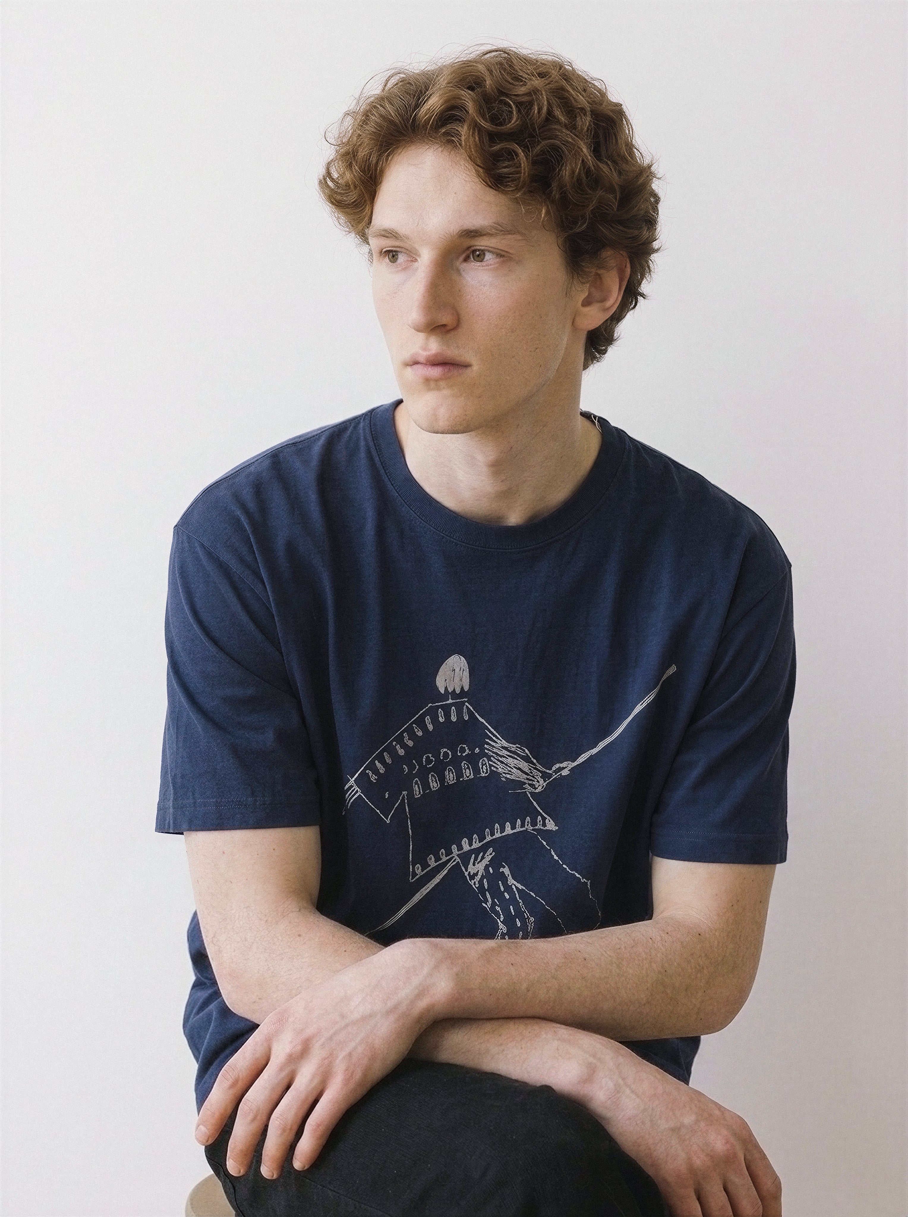 A male model sitting and looking away, demonstrating the relaxed drape of a vintage navy oversized streetwear t-shirt with a balancing figure graphic.
