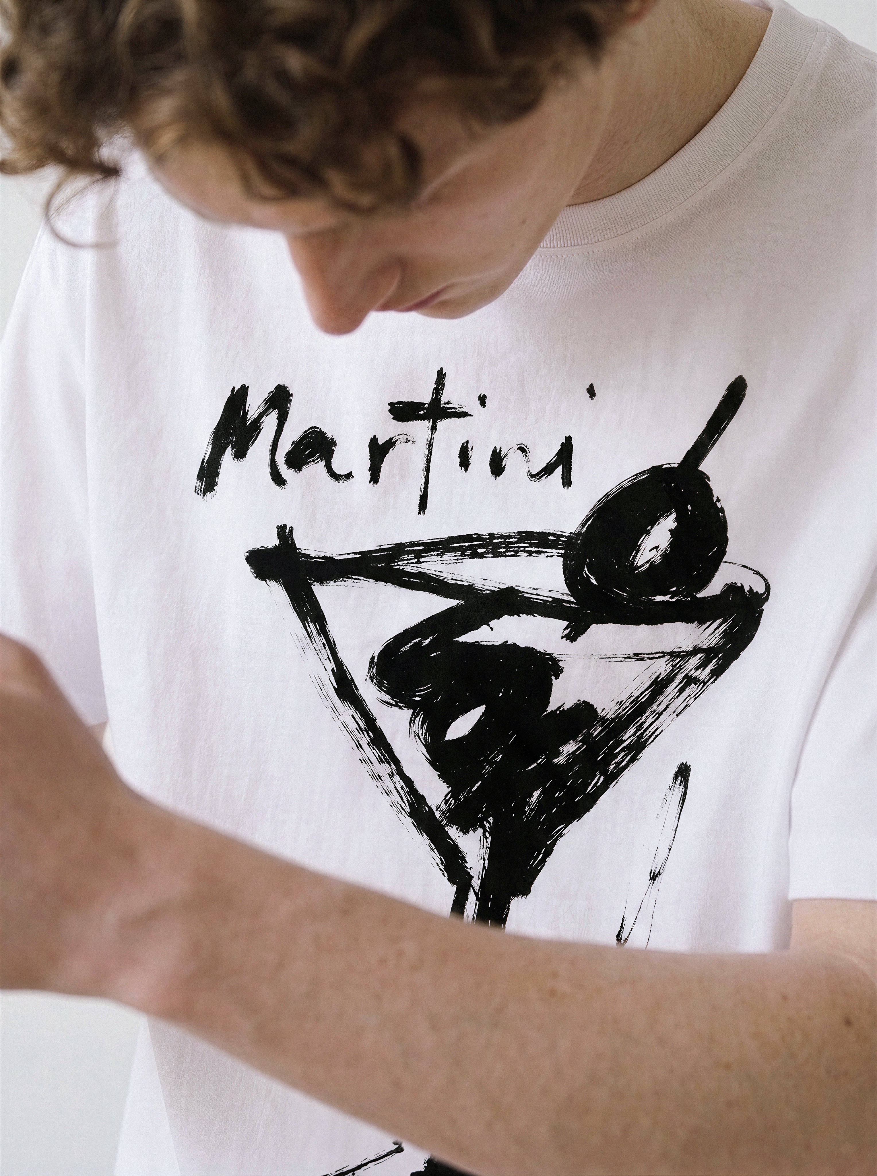 Close-up view of a model looking down, emphasizing the bold black martini glass and handwritten typography line art on a white t-shirt.