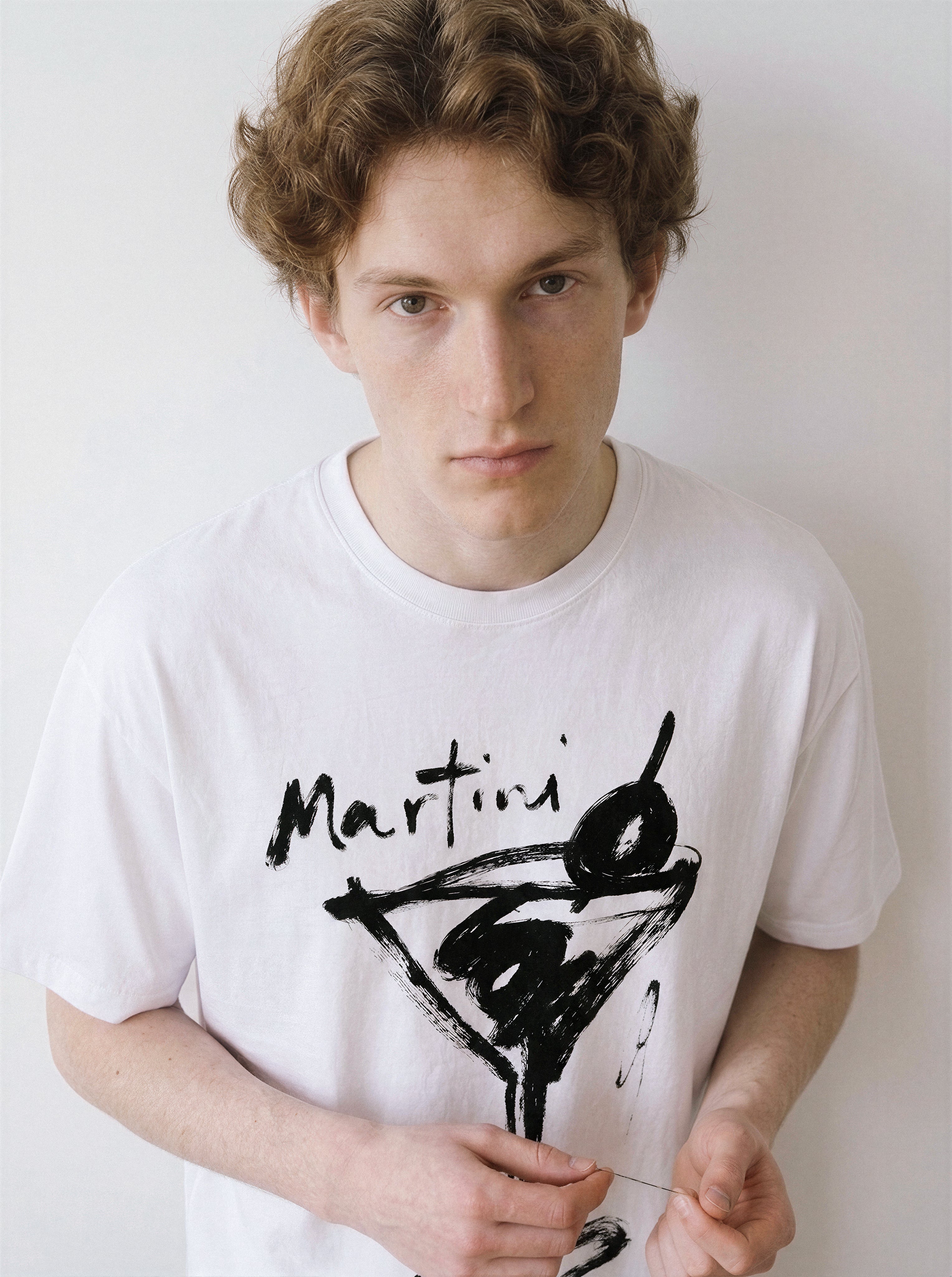 Front view of a male model looking directly at the camera, wearing a clean white graphic tee highlighting an expressive martini glass line art illustration.