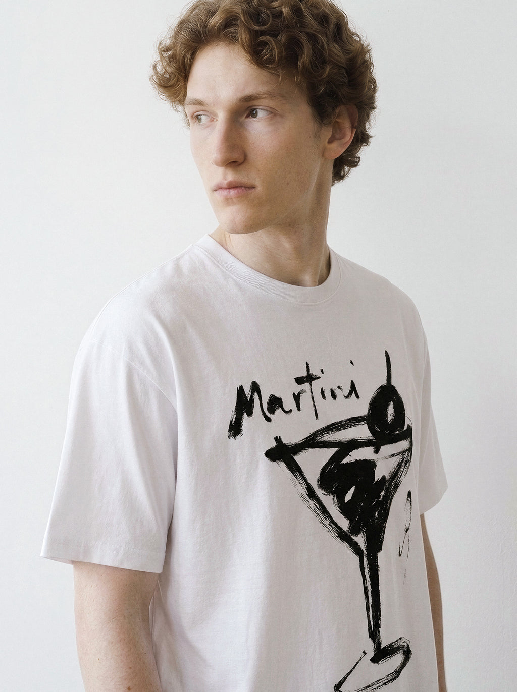 Front portrait of a male model looking away, wearing a white t-shirt featuring a minimalist brush-stroke martini glass line art graphic.