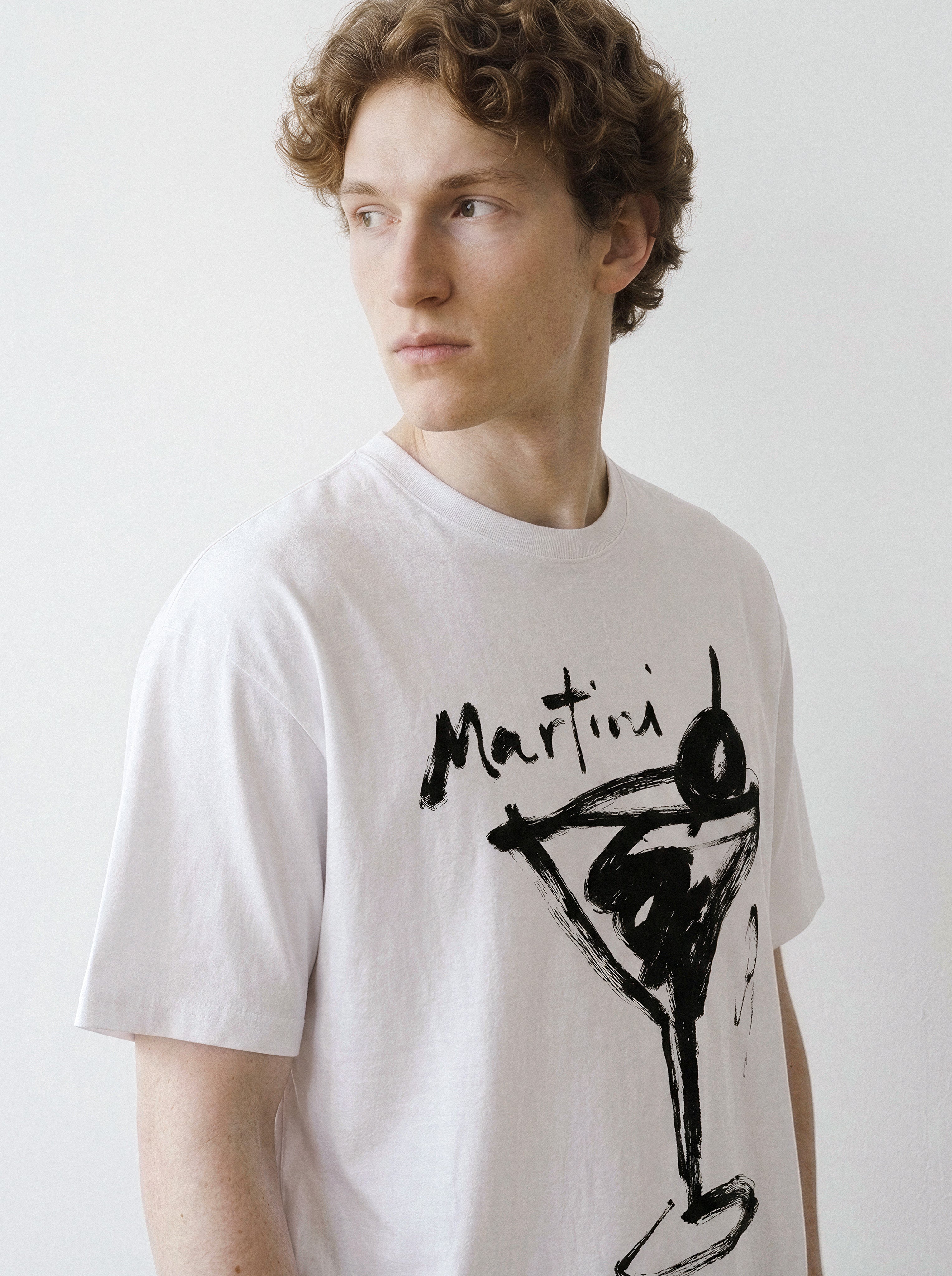 Front portrait of a male model looking away, wearing a white t-shirt featuring a minimalist brush-stroke martini glass line art graphic.