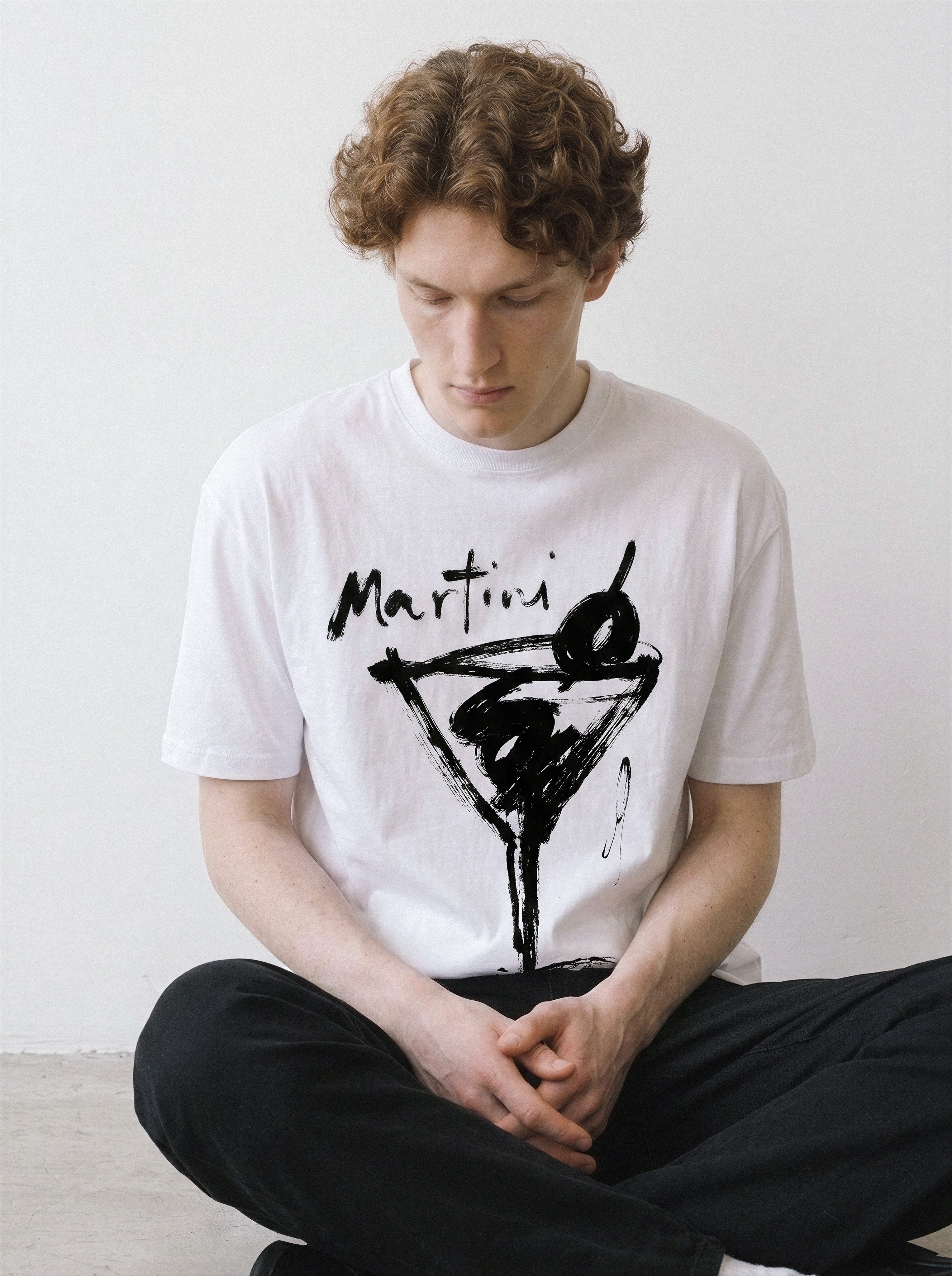A young man with curly brown hair sits cross-legged against a plain white wall, wearing a white crewneck t-shirt with a black hand-drawn martini glass and 'Martini' text graphic, paired with black pants.