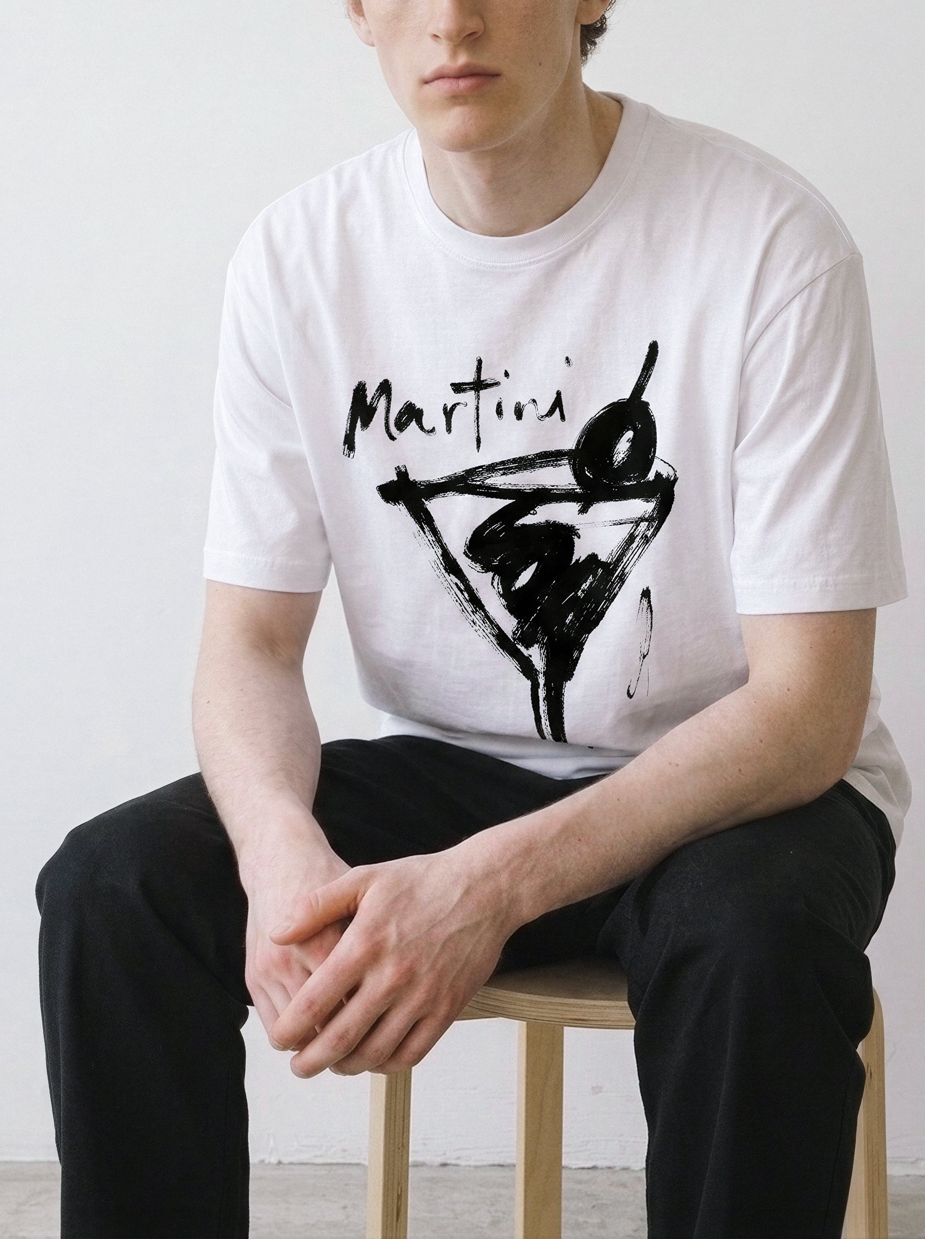 Model sitting on a stool with hands on knees, demonstrating the drape of a white t-shirt with a rough brush-stroke martini line art print.