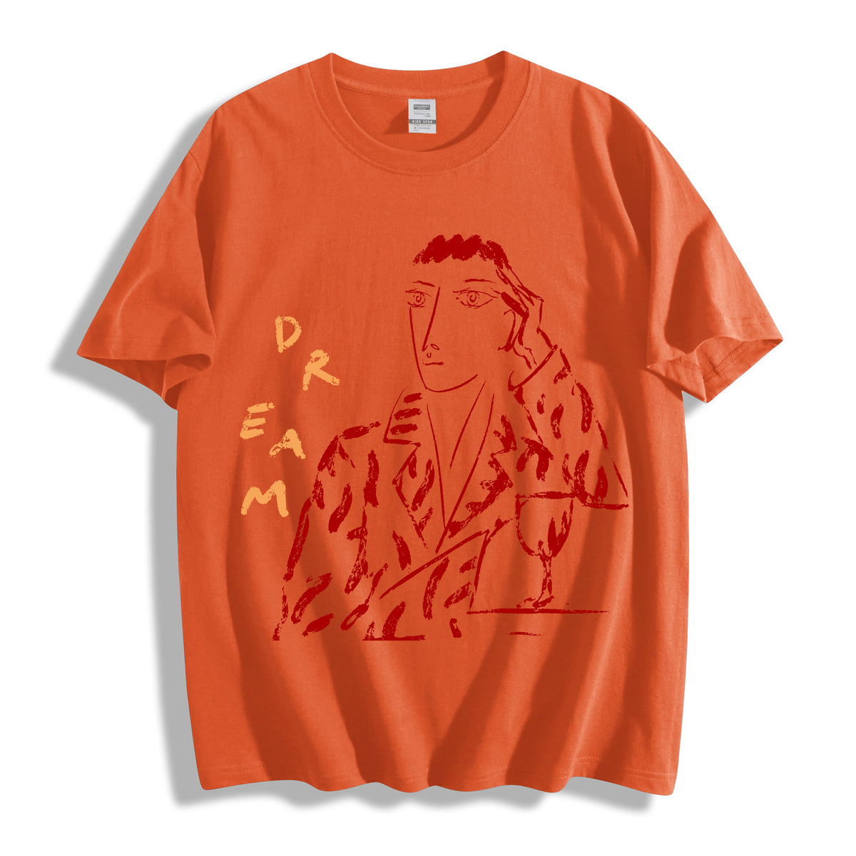 An orange short-sleeve crewneck t-shirt featuring a red graphic print of a stylized figure and the word 'DREAM' in vertical letters on the front, displayed against a clean white background.
